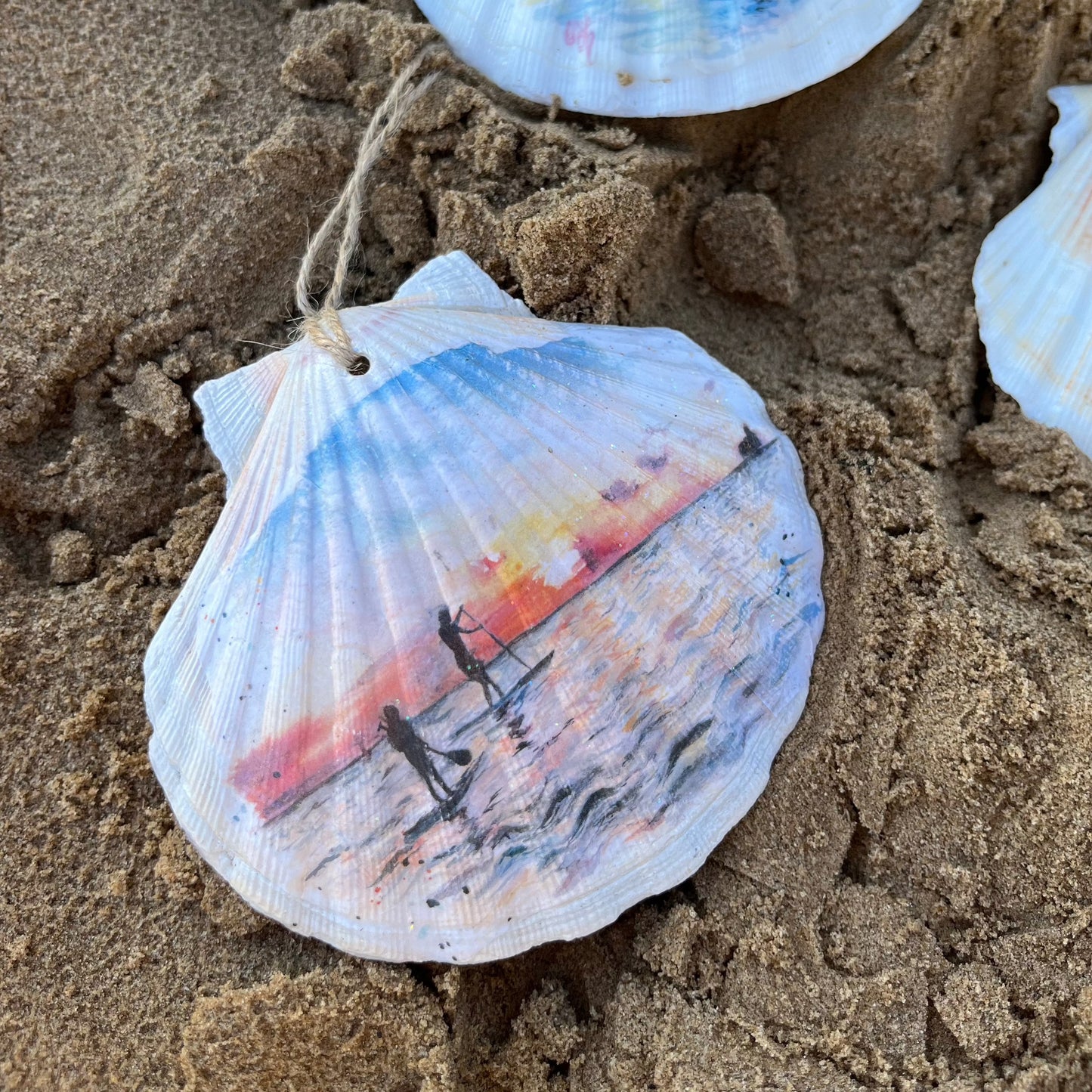 A hanging decoupaged shell featuring watercolour artwork of paddleboarders on Cleethorpes Beach by Eve Leoni Art and Jollpotz.