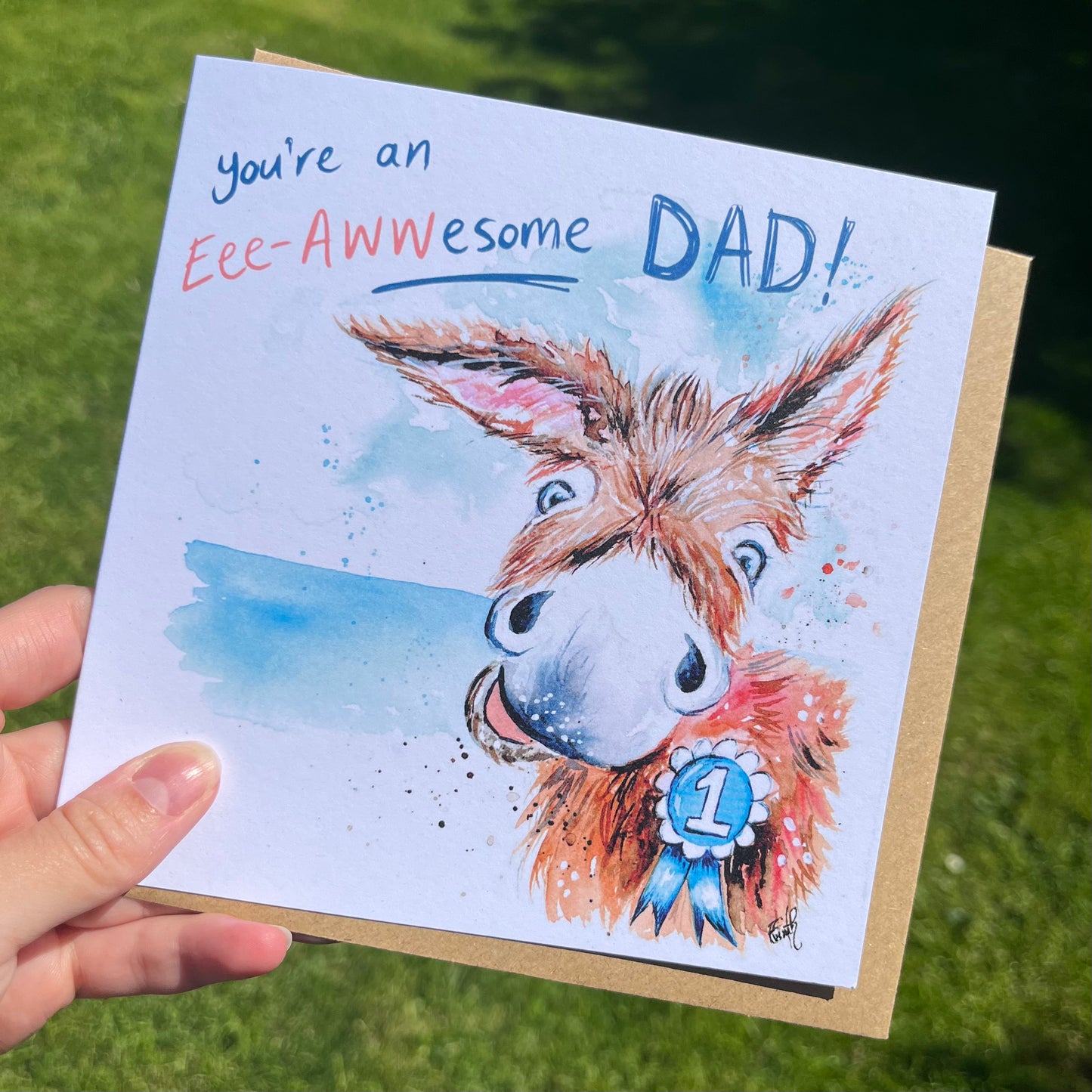 A greetings card featuring an excited donkey wearing a rosette, with the writing, 'You're an Eee- Awwesome Dad!', illustrated by local artist Eve Leoni Smith.
