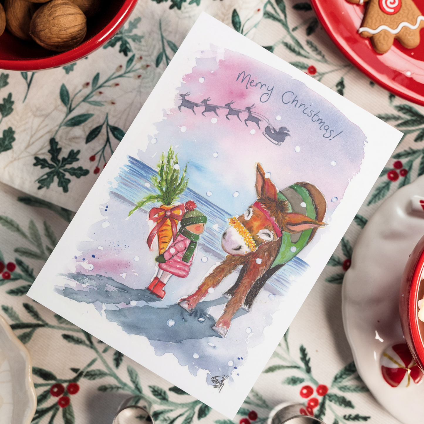 A watercolour Christmas Card featuring a young girl waiting to gift a large carrot to Dudley the Donkey for Christmas on Cleethorpes beach. Painted by Grimsby artist, Eve Leoni Smith.