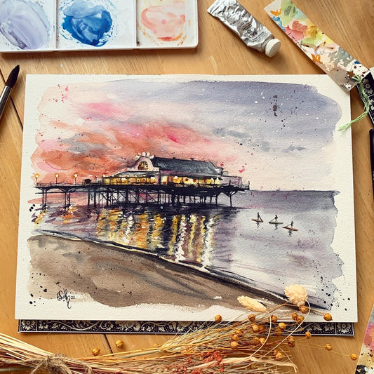 An original watercolour print design by Cleethorpes artist, Eve Leoni Smith, featuring the Cleethorpes pier at sunset with some stand up paddleboarders enjoying the evening water. 