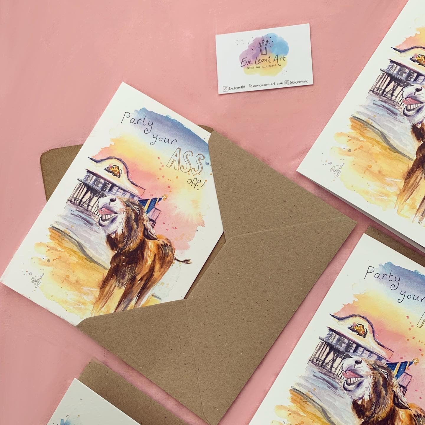 Greetings cards with brown kraft envelopes laid on a pink background, featuring local watercolour artwork of a Cleethorpes donkey wearing a party hat in front of Cleethorpes Pier. The card says, 'Party your ass off!'