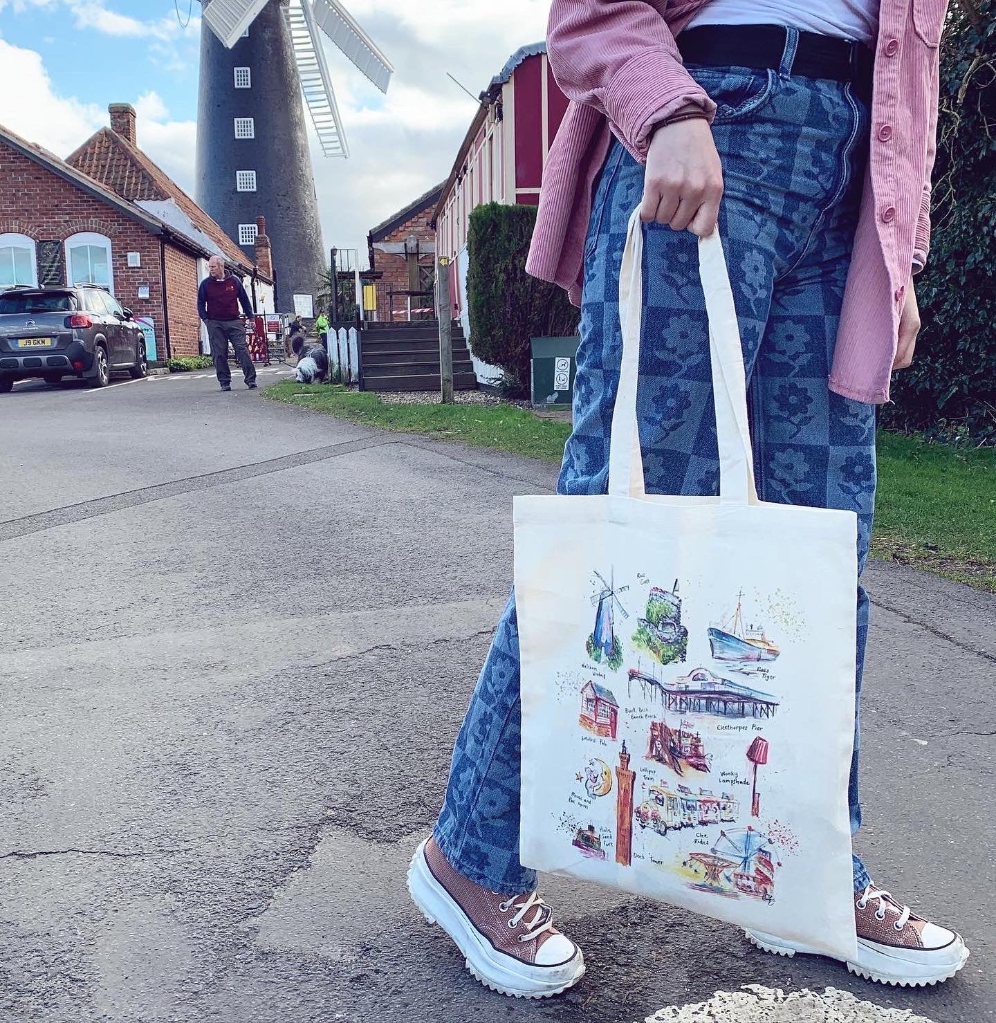 Model holding an Organic tote bag in front of the Waltham Windmill, featuring watercolour illustrations of landmarks in Grimsby and Cleethorpes, designed by local artist, Eve Leoni Smith.