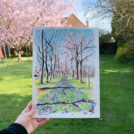 An original watercolour painting being held up in front of a blossom tree. The painting features the trees in People's Park, Grimsby, during the spring, painted by Eve Leoni Art. 