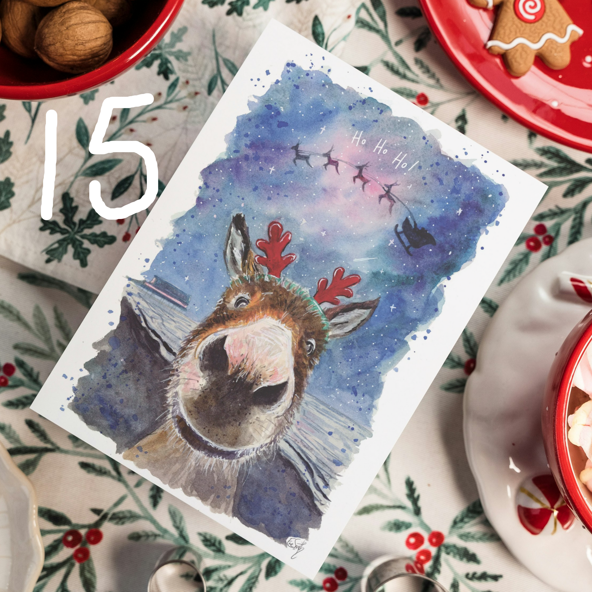 A Christmas card featuring a watercolour illustration of a smiling donkey popping up from the bottom of the page, he is on Cleethorpes Beach and wearing some dress-up Rudolph antlers. Santa and his reindeers are flying in the sky above and text reads 'Ho Ho Ho!'. Painted by Eve Leoni Art.