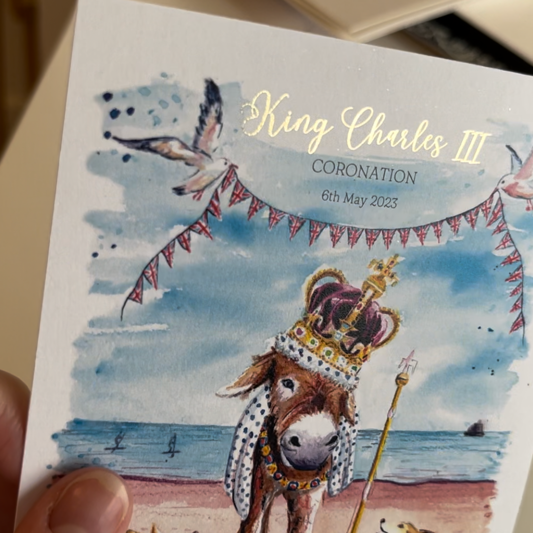 Limited edition postcards featuring a donkey wearing the crown jewels on Cleethorpes beach, surrounded by royal corgis to commemorate the coronation of King Charles III. His Majesties name is written in gold foil and shines in the light. 