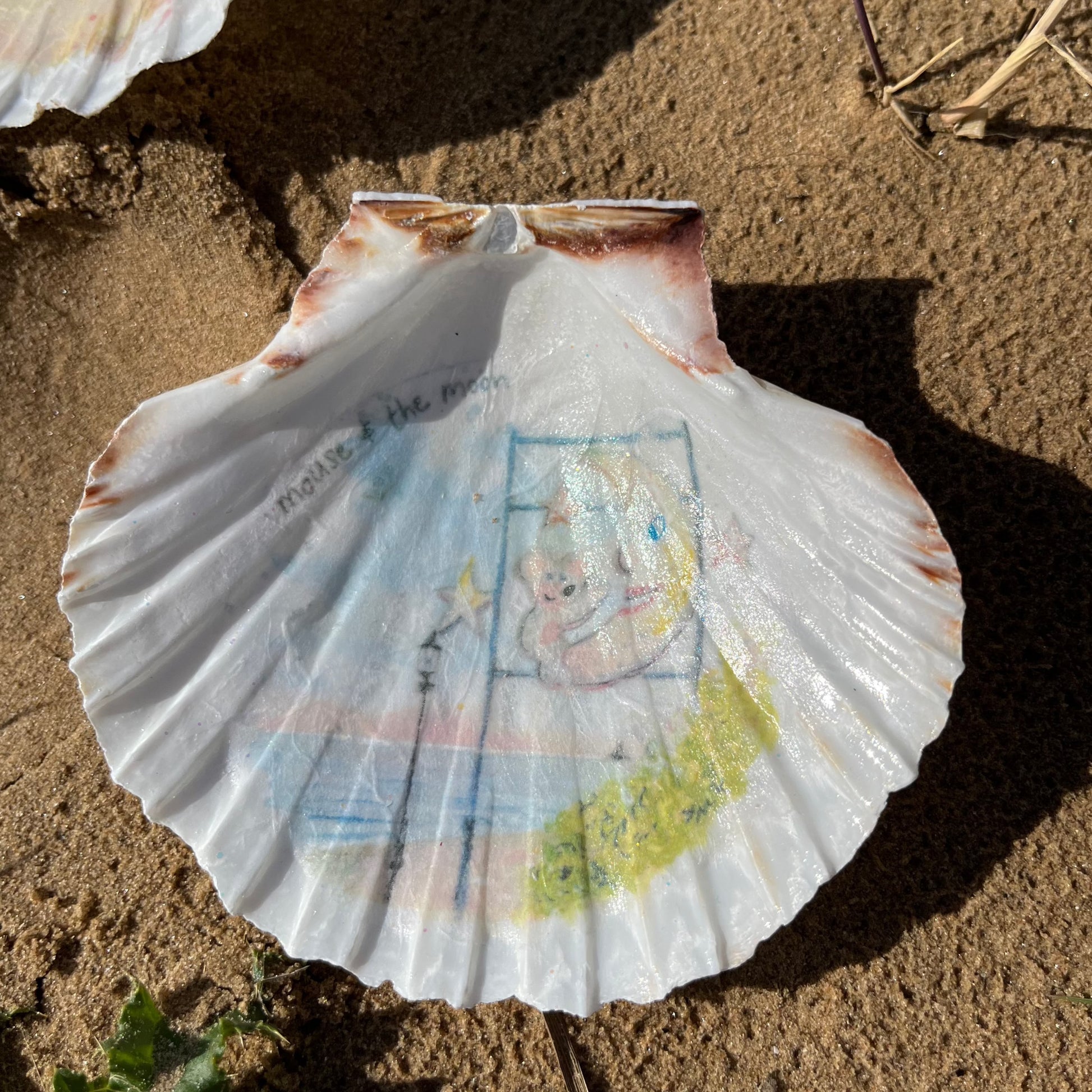 A decoupaged shell on Cleethorpes beach, featuring watercolour artwork of the Mouse and the Moon by Eve Leoni Smith and Jollypotz.