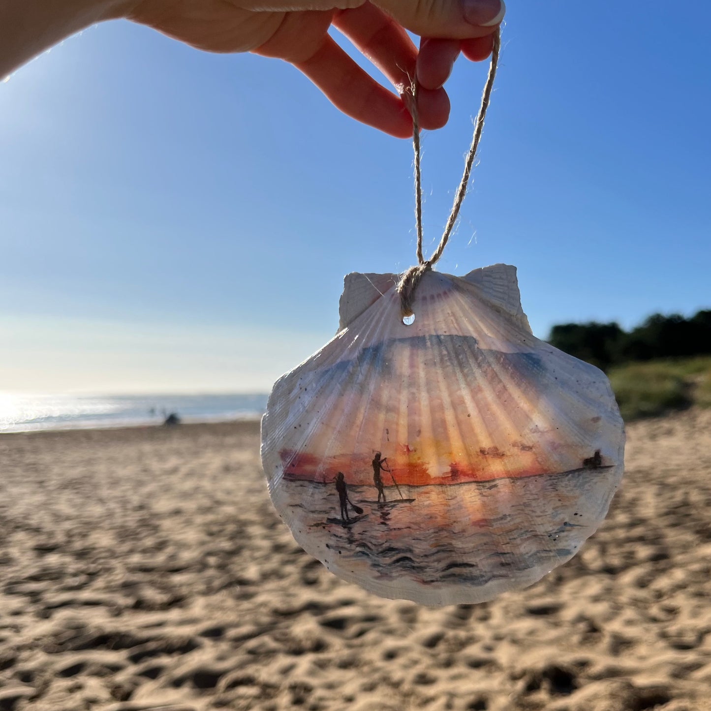 A hanging decoupaged shell featuring watercolour artwork of paddleboarders on Cleethorpes Beach by Eve Leoni Art and Jollpotz.