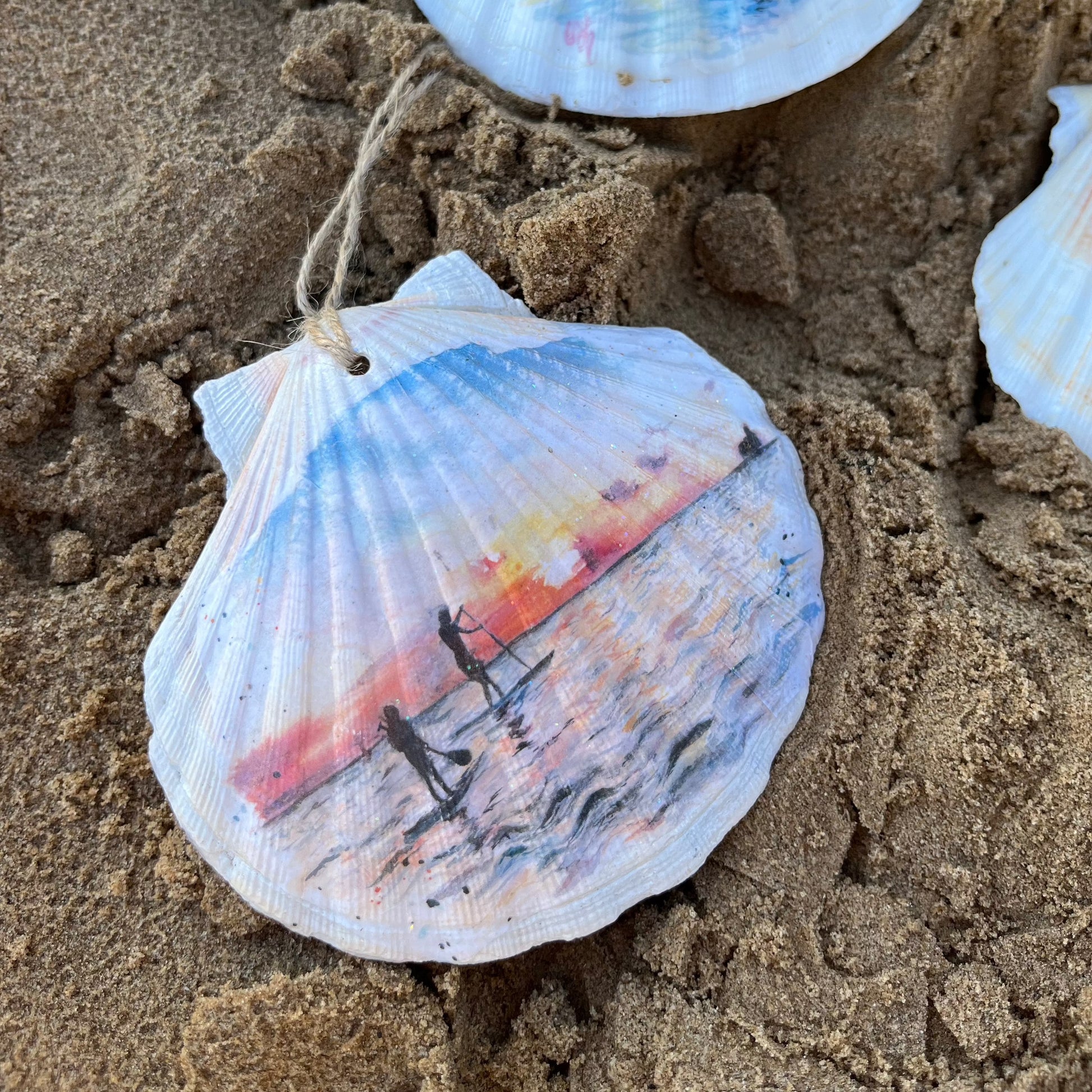 A hanging decoupaged shell featuring watercolour artwork of paddleboarders on Cleethorpes Beach by Eve Leoni Art and Jollpotz.