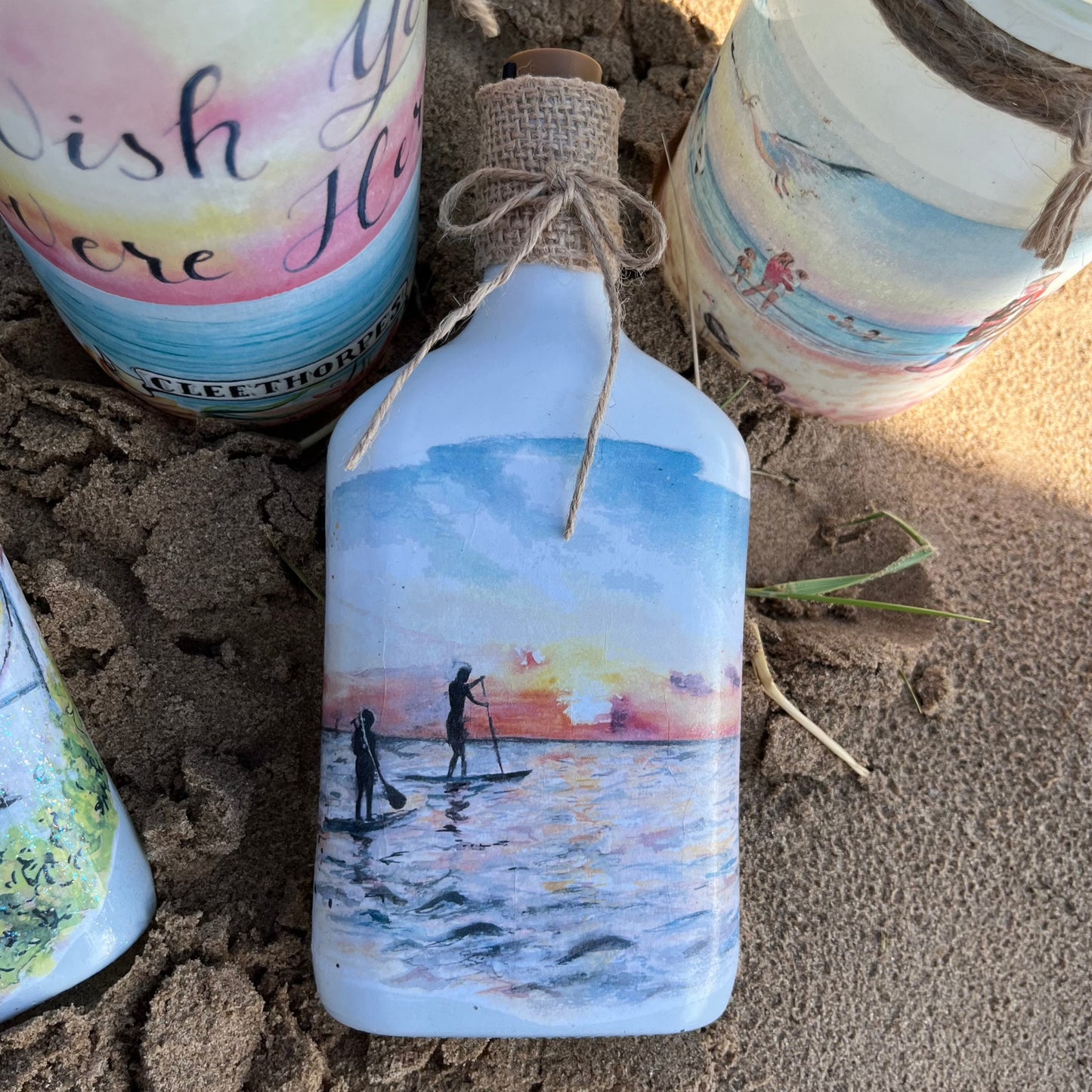 A decoupaged jar on Cleethorpes beach, featuring watercolour artwork of paddleboarders at sunrise by Eve Leoni Smith and Jollypotz.
