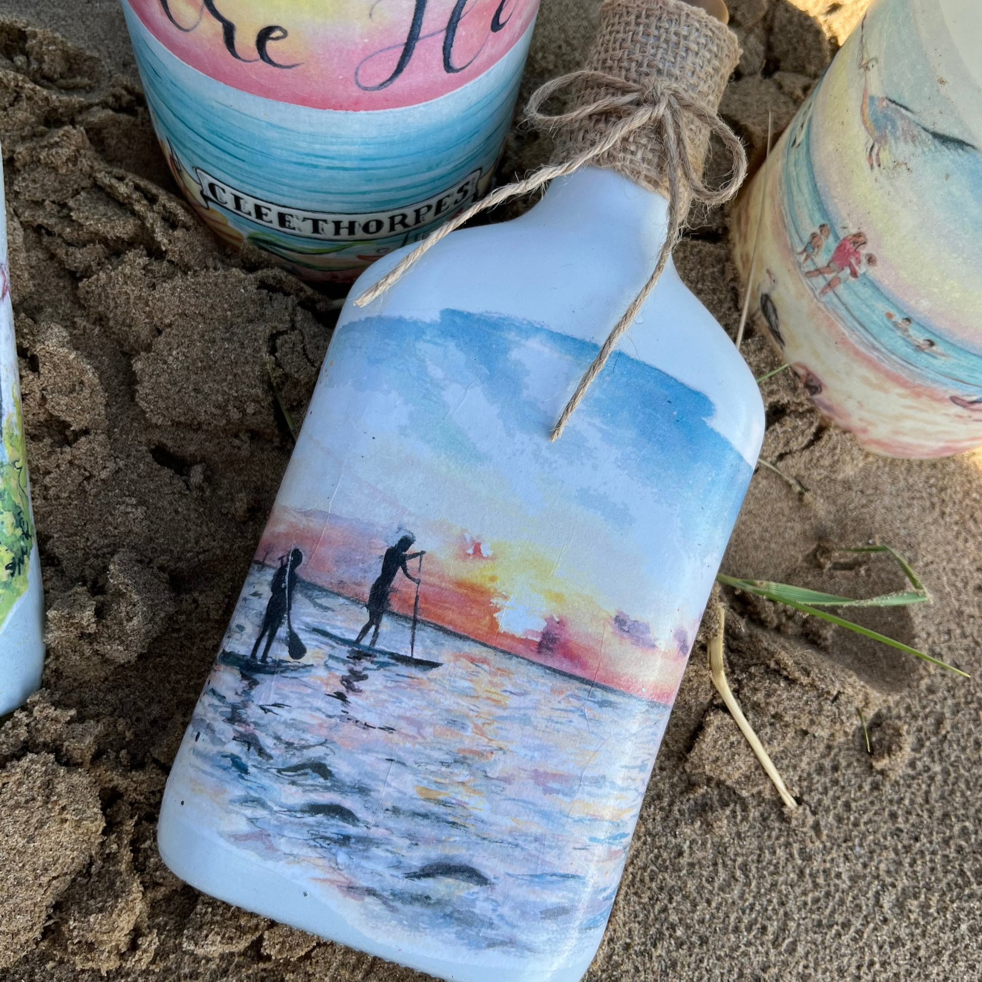 A decoupaged jar on Cleethorpes beach, featuring watercolour artwork of paddleboarders at sunrise by Eve Leoni Smith and Jollypotz.