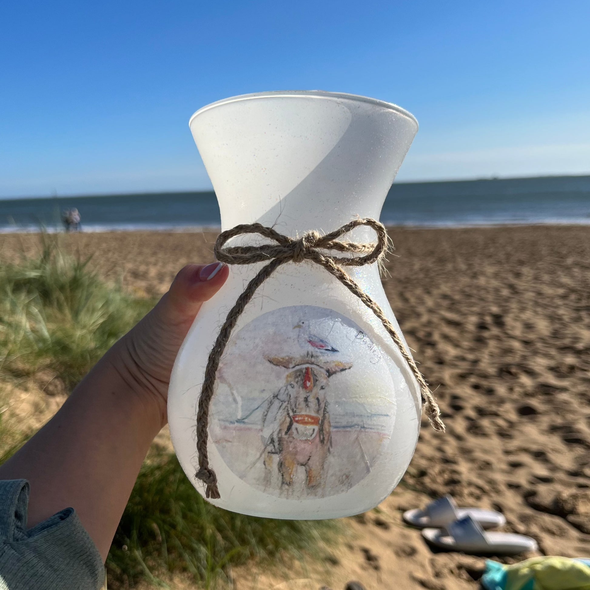 A decoupaged vase on Cleethorpes beach, featuring watercolour artwork of Dudley the Donkey by Eve Leoni Smith and Jollypotz.