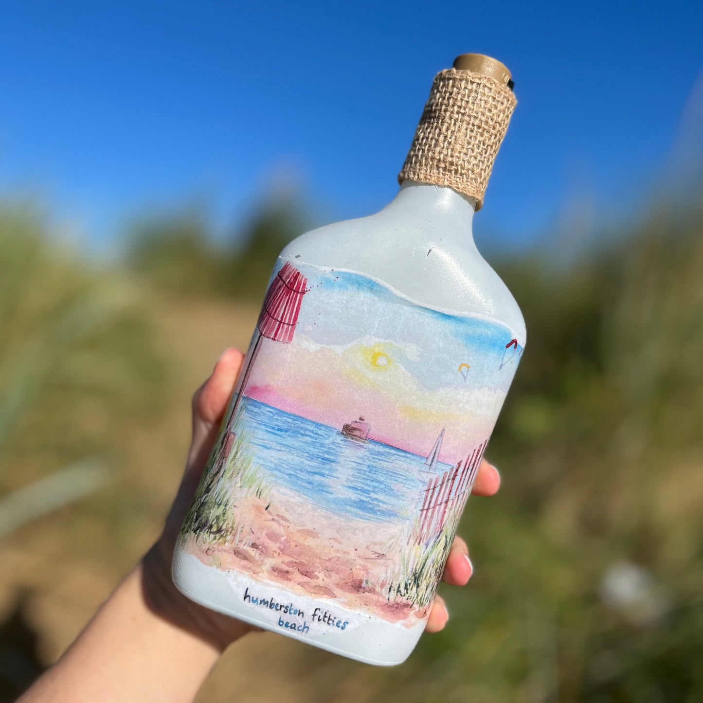 A decoupaged bottle on Cleethorpes beach, featuring watercolour artwork of the Humberston Fitties by Eve Leoni Smith and Jollypotz.