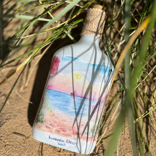 A decoupaged bottle on Cleethorpes beach, featuring watercolour artwork of the Humberston Fitties by Eve Leoni Smith and Jollypotz.