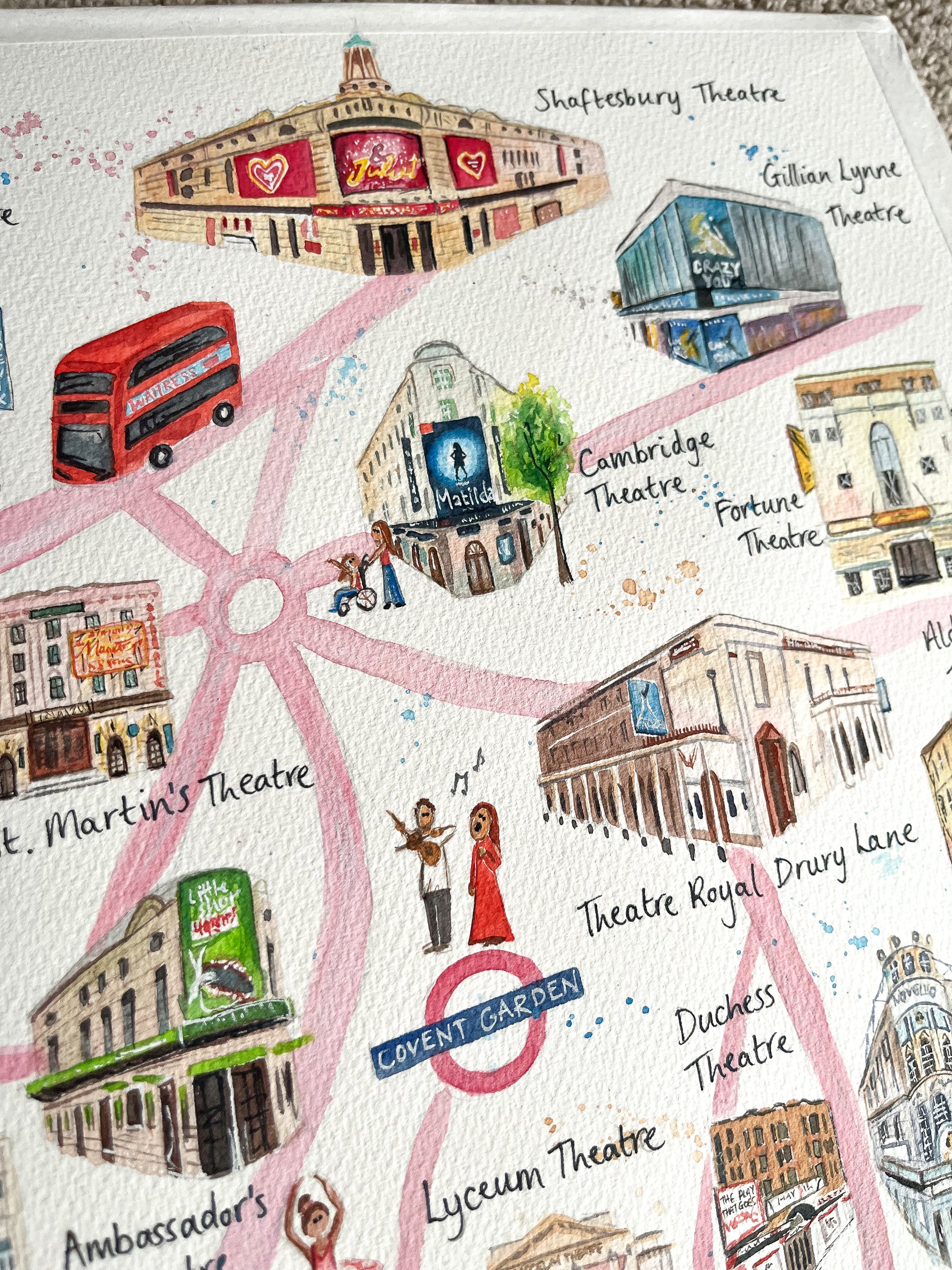 A close up photo of a watercolour map of the West End by artist, Eve Leoni Smith. It includes theatre illustrations of the Shaftesbury Theatre, Theatre Royal Drury Lane and Cambridge Theatre. 