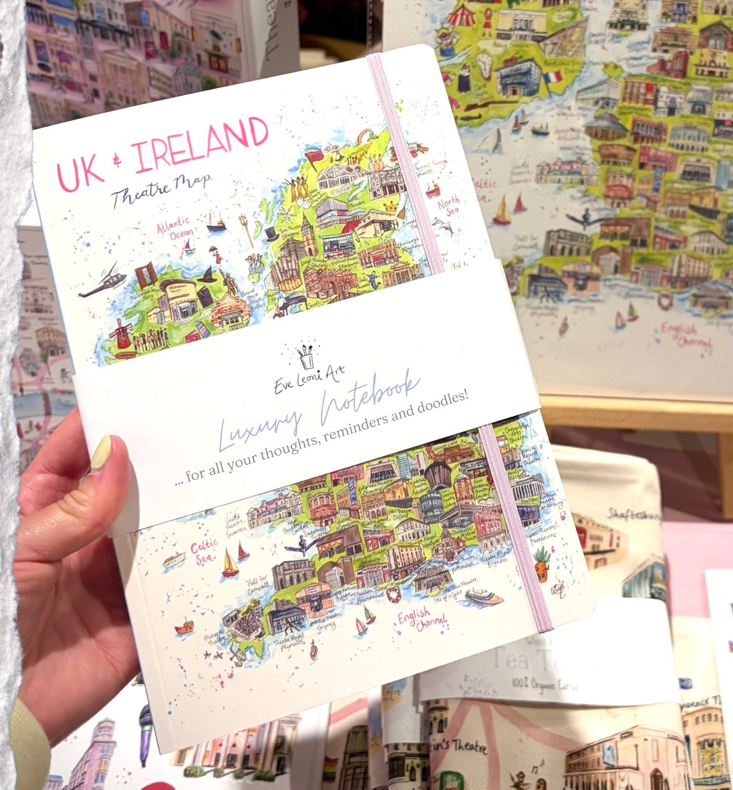 A notebook with pink elastic showing a watercolour illustration of a UK and Ireland map featuring regional theatres and musical venues by Eve Leoni Art. 