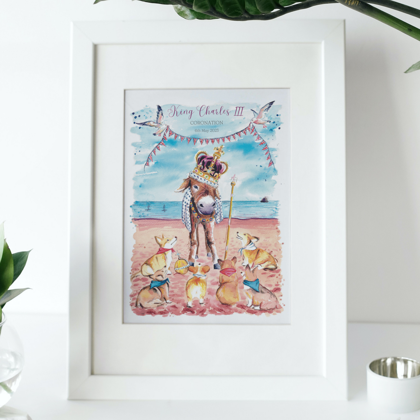 A limited edition signed print to commemorate King Charles III Coronation, featuring a donkey in the crown jewels on Cleethorpes beach surrounded by some royal corgis. Painted in watercolours by Eve Leoni Smith and framed in a white frame.